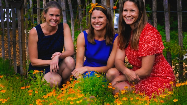 Tre jenter i fargerike sommerkjoler sitter i en blomstereng og ønsker velkommen