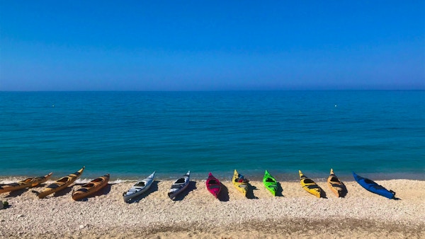 Elleve kajakker i forskjellig farge ligger på rad og rekke på stranden foran det turkise havet