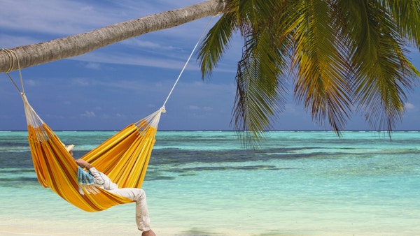 En ung mann iført stråhatt slapper av i en oransje hengekøye som er hengt på et kokosnøttpalm. Den hvite sandstranden og fargen på vannet er turkis og er en idyllisk og unik beliggenhet på Maldivene.
