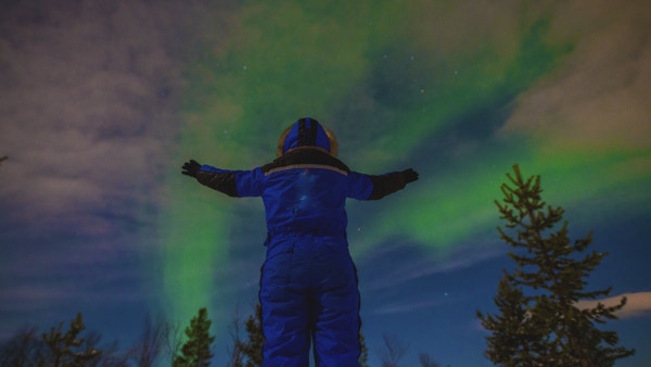 Turist med hette som ser på nordlyset på nattehimmelen i vinterlandskap, Kirkenes, Finnmark, Norge