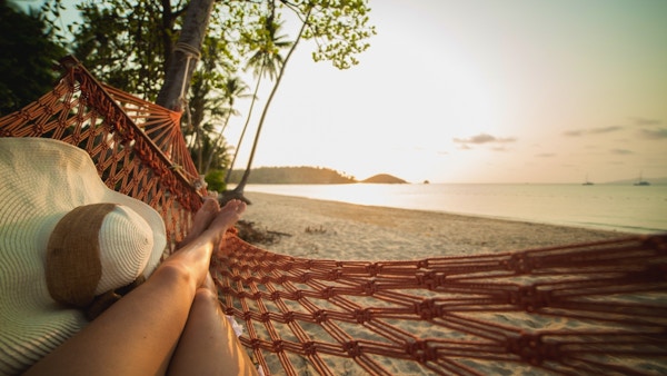 Beina til kvinne i hengekøye med solhatt og utsikt til strand, hav og palmer.