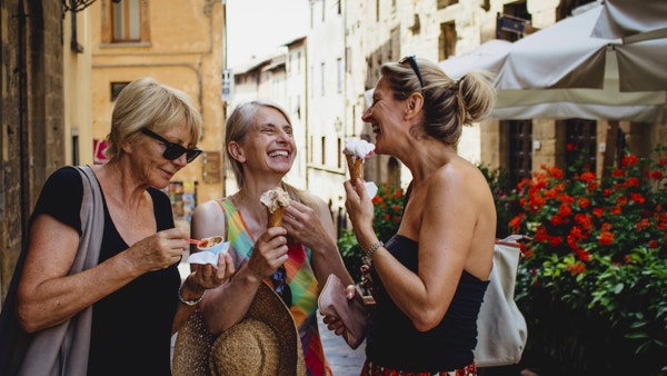 Tre venninner som står og spiser italiensk iskrem mens de står i en gate i Toscana om sommeren. De smiler og vender seg mot hverandre mens de nyter ferien.