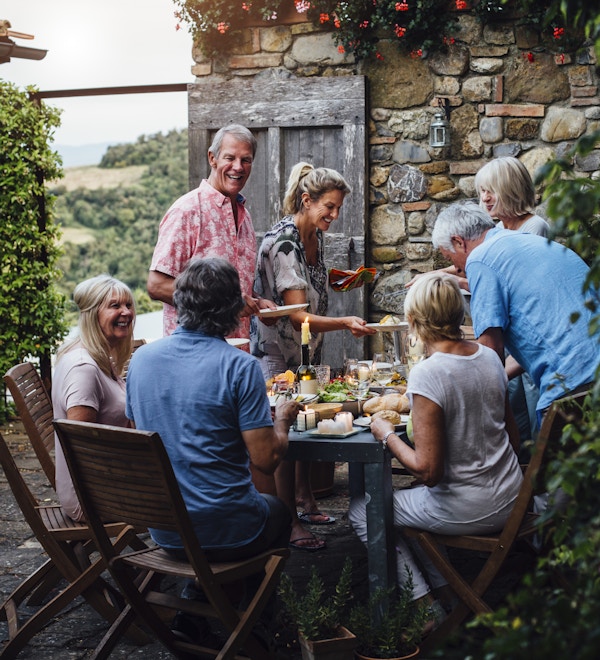 En gruppe voksne venner sitter rundt et utendørs spisebord, spiser og drikker. De snakker alle lykkelig og nyter hverandres selskap. Bildet er tatt i Toscana, Italia.