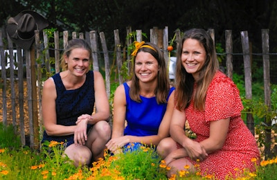 Tre jenter i fargerike sommerkjoler sitter i en blomstereng og ønsker velkommen