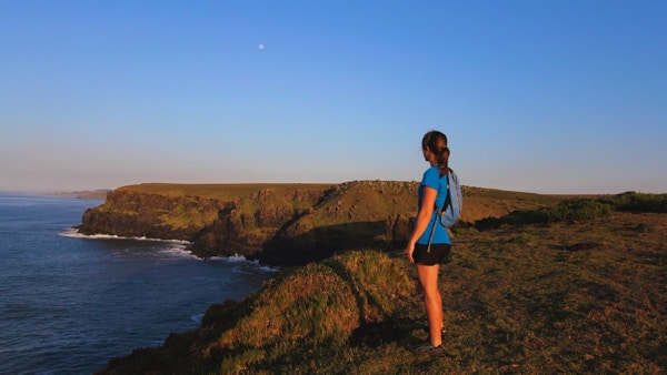 kvinne med ryggsekk står med ryggen til og ser utover havet fra ei klippe