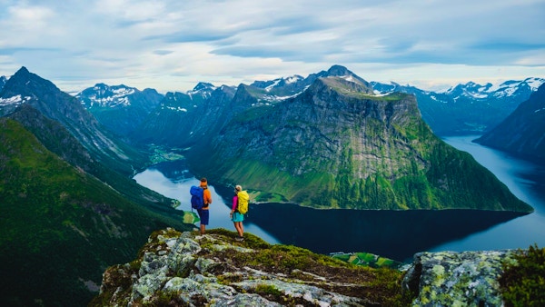 2 mennesker med turklør og tursekk på ryggem på toppen av fjellet og ser ut over Hjørundfjorden