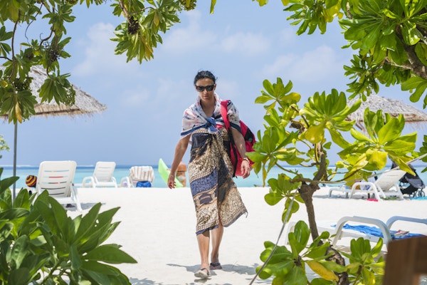 Vindusvisning av vakker ung kvinne som bærer en sarong og går på en bikinistrand på Gulhi-øya, Maldivene. I bakgrunnen står solstoler med solbeskyttende parasoller, Det indiske hav og skyer på blå himmel ligger i bakgrunnen. Gulhi-øya ligger ikke langt fra Maafushi-øya på Maldivene, og er åpen for utlendinger.