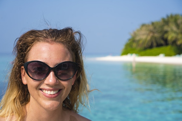Portrett av en ung kvinnelig turist med solbriller, glad og smilende på en sandstrand med turkis vann, blå himmel og grønn vegetasjon i bakgrunnen. Bildet er tatt i Det indiske hav ikke langt fra Maafushi Island på Maldivene.