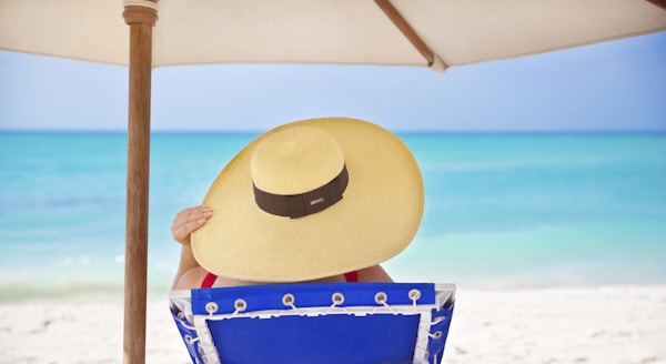 En uidentifiserbar kvinne som slapper av og har på seg badedrakt og solhatt mens hun leser en bok og slapper av på en blå og hvit stripet strandstol. Mens hun sitter komfortabelt, ser hun på havutsikten på en hvit sandstrandferie ved Det karibiske hav ved Playa del Carmen, nær Cancun, Riviera Maya, Mexico.