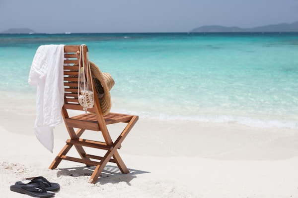 tom teak stol med stråhatt, hvit skjorte, sandaler og en øl på karibiske stranden