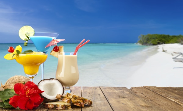 Pina colada, fersken daiquiri og blå curaçao cocktailer på et trebord med tropisk strandbakgrunn. Disse drinkene tilbys mest på tropiske feriedestinasjoner. Ved bunnen av glassene er en halv og en hel kokosnøtt, tre skiver ananas og en rød hibiskusblomst. Forgrunnskomposisjonen er til venstre for rammen og etterlater et nyttig kopierom midt til høyre.