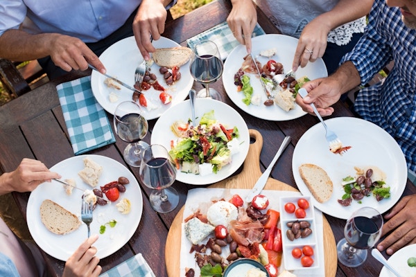 Bord fotografert ovenfra med folk som spiser lunsj, brød, oliven, ost, salat og rødvin.