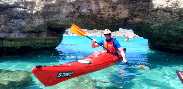 Mann med hatt og solbriller padler en rød kajakk på turkist hav under en bro av kalkstein