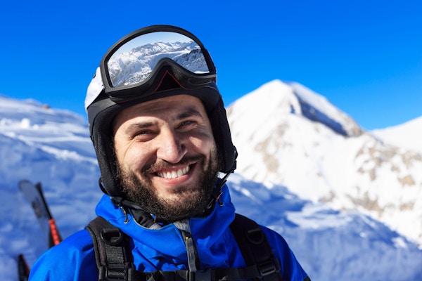 Smilende ung mann som vandrer på skianlegg. Med skiklær, vernebriller, hjelm og ryggsekk. Ser på kamera. I bakgrunnen en fjelltopp med snø. Fokus på forgrunnen.