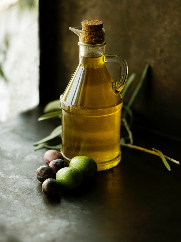 En glasskrukke med olivenolje står på et bord, korket og med noen oliven foran
