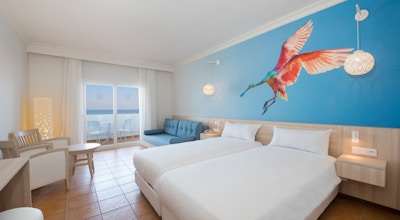 Hotellrom med to senger med hvitt sengeteppe samt en stor fargerik fugl på tapet bak. Stol og sittegruppe