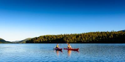 Landskapsbilde av to mennesker i hver sin kajakk som padler på innsjøen med blå himmel og skog i bakgrunnen