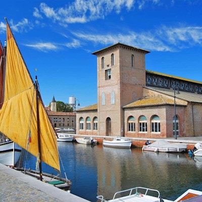 Langs bryggekanten med fortøyde båter i flere størrelser, noen med gule og røde seil og en gammel bygning i bakgrunnen på andre siden av kanalen