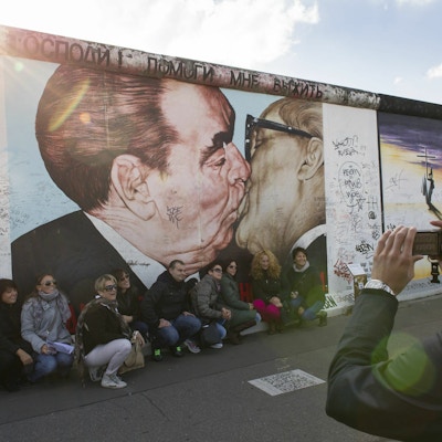 En gruppe mennesker poserer foran berlinmuren og broderkysset mens de blir tatt bilde av