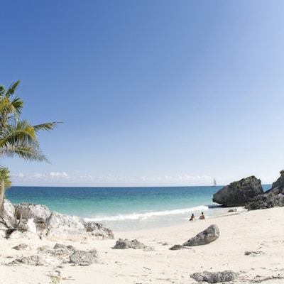 En hvit strand med palmer og to mennesker som sitter i vannkanten