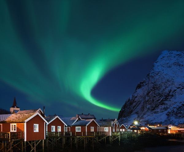 Nydelig form på nordlys (Aurora borealis) over tradisjonelle norske fiskehytter (rorbuer) og snøhvit fjell.