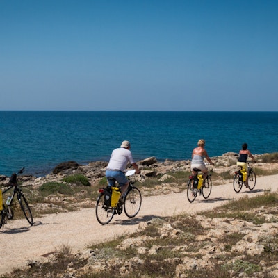 Tre mennesker sykler forbi en parkert sykkel på grusvei langs havet på vei mot Capilungo, Puglia