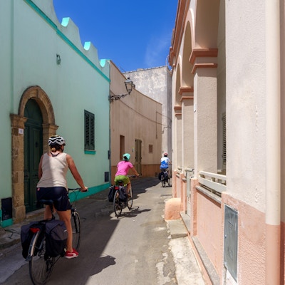 Tre personer sykler på en smal vei mellom pastellfargede, lave hus i Tuglie
