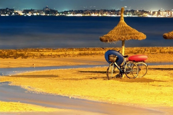 To sykler står parkert under en stråparasoll på en strand med en blå og en rød badering hengt  over styret