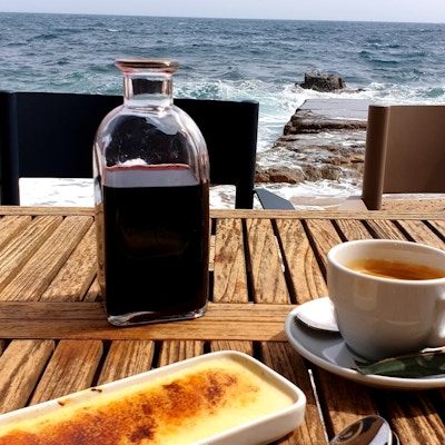 Desserten Crema Catalana på en tallerken med en karaffel rødvin og en kaffekopp på bordet. Bestikk. Utsikt utover havet.