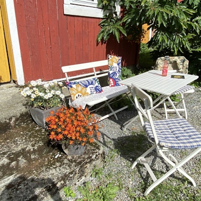 Trebygning med stoler og bord med rutete pute på samt blomster i krukke
