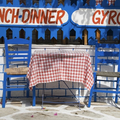 To blå stoler og et bord med rutete duk foran et skilt med lunch, dinner og gyros på vinduet