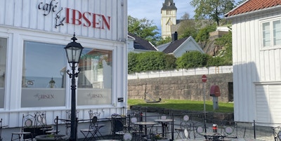 Spiseområde med bord og stoler utenfor Café Ibsen i Girmstad, med blå himmel og Grimstad kirke i bakgrunnen