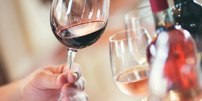 Nærbilde av ugjenkjennelige voksne som holder glass vin og utfører visuell undersøkelse. Evaluering av utseende, farge, klarhet, rester på glasset. Det er vinflasker og andre vinprøver i bakgrunnen.