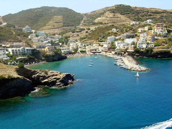 Blått hav, klipper, havn og mange båter som ligger foran hvit bebyggelse på Kreta