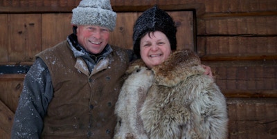 En mann og ei dame ikledd vinterklær og pels ønsker velkommen til fjells foran ei tredør