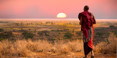 Masai-mann, med tradisjonelle tepper, har utsikt over Serengeti i Tanzania når den fargerike solnedgangen fyller himmelen. Villt gress i marken.