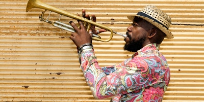 En kjent profesjonell cubansk musiker med skjegg, iført en Panama-hatt, står mot en lukket gul butikklukker, spiller en trompet utendørs, Havana, Cuba, 50 megapiksler bilde