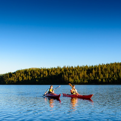 Landskapsbilde av to mennesker i hver sin kajakk som padler på innsjøen med blå himmel og skog i bakgrunnen