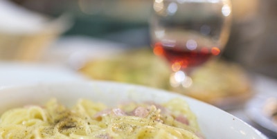 Nærbilde av en bolle med spaghetti a la carbonara. Et glass rødvin og brød er synlige i bakgrunnen.