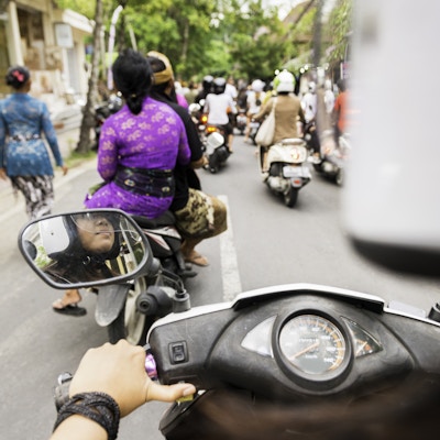 Dette over skulderfrie arkivfotografiet er fra motorsykkel bakre passasjer synspunkt. En 18 år gammel indonesisk kvinne kjører kjøretøyet. Hjelmen hennes er ute av fokus i forgrunnen og gir kopiplass. Veien videre i Ubud, Bali, er full av andre motorscootere og fotgjengere som har på seg fargerike, tradisjonelle balinesiske klær i løpet av sent på ettermiddagen. Fotografert med et Nikon D800 DSLR-kamera.