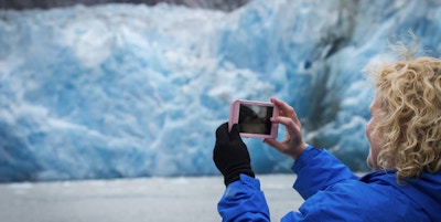 Blond kaukasisk kvinne i 40-årene fotograferer den blå isen på North Sawyer Glacier med smarttelefonen sin som passasjer på et cruiseskip, Tracy Arm Fjord, Juneau, Alaska