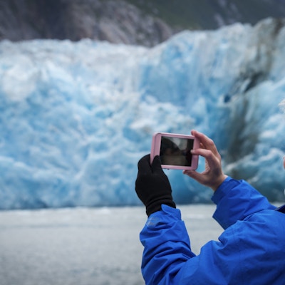 Blond kaukasisk kvinne i 40-årene fotograferer den blå isen på North Sawyer Glacier med smarttelefonen sin som passasjer på et cruiseskip, Tracy Arm Fjord, Juneau, Alaska