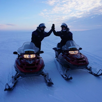 To personer kjører hver sin snøskuter i solnedgang