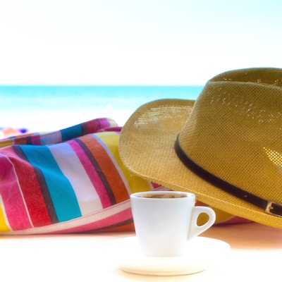 Et nærbilde av en hvit kopp espresso, en solhatt og en stripet strandbag på et hvitt bord; i den uskarpe bakgrunnen er et turkis havlanskap og lys himmel med bare en blåtone. Bilde tatt i Italia