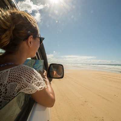 Kvinne i bil leser kart og ser ut på sanstrand, hav og horisont.