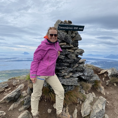 Kvinne i lilla jakke og turutstyr står på toppen av et fjell i Norge