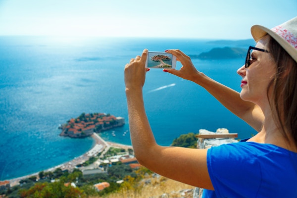 En kvinne med solhatt som tar bilder ved byen Sveti Stefan i Montenegro, Balkan