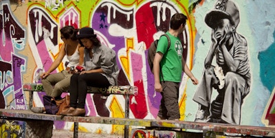 To ungdommer sitter på en benk foran en vegg med grafitti mens en mann går bak dem og ser på veggen