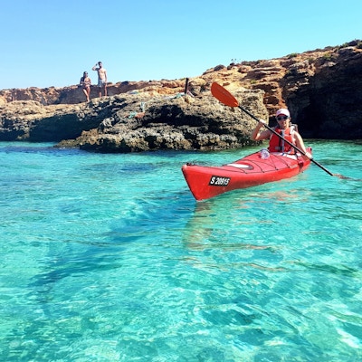 En dame padler en rød kajakk på turkist hav med kalksteinsformasjoner i bakgrunner, to mennesker ser på henne padle fra land