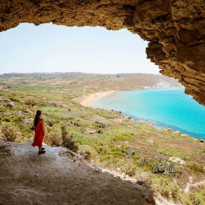 En dame i rød kjole står i Mixta grotte og ser utover landskapet med havet og den røde stranden der nede
